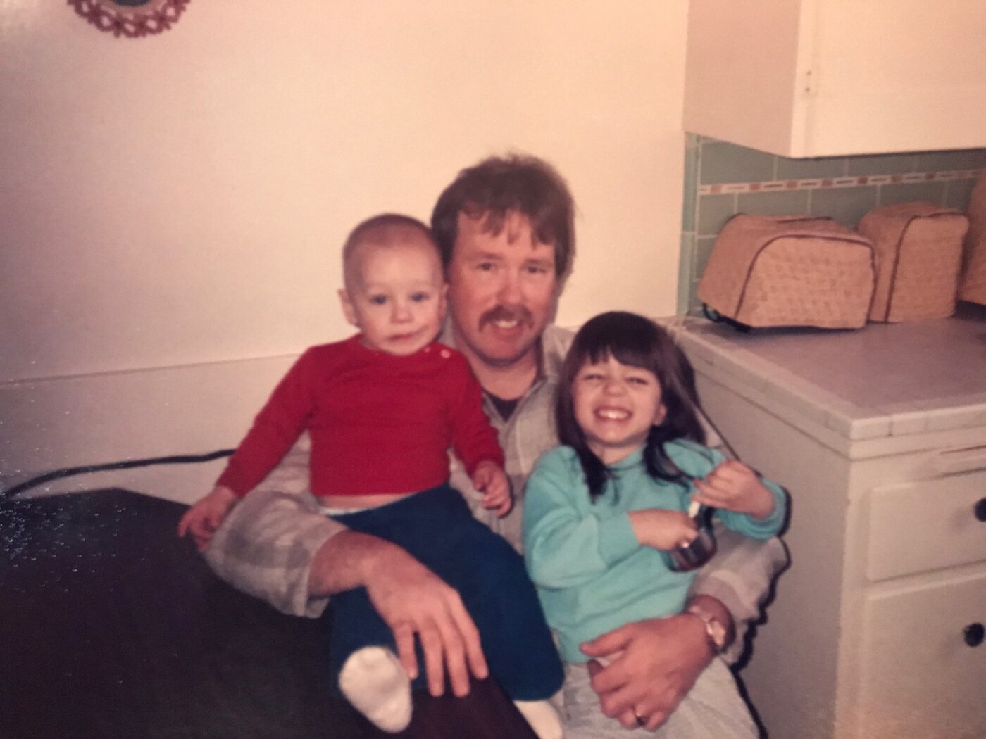 Robert Fitch sitting on the floor holding his two young children, his gold 25-year service watch on his wrist